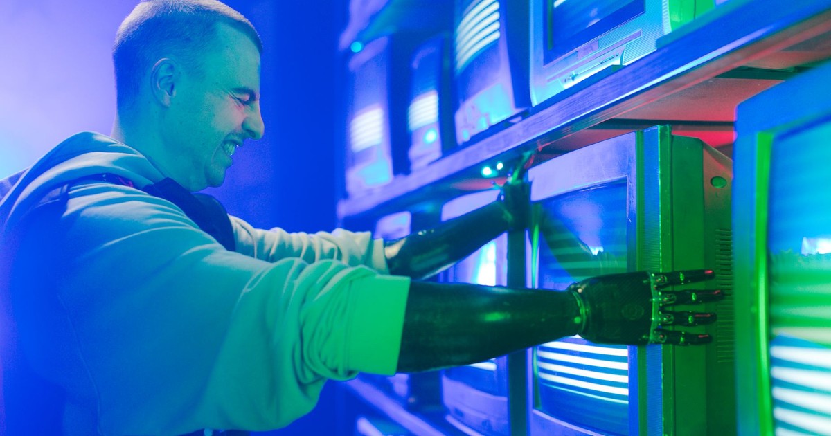 Smiling man with prosthetic arm adjusts retro TVs in a neon-lit setting.