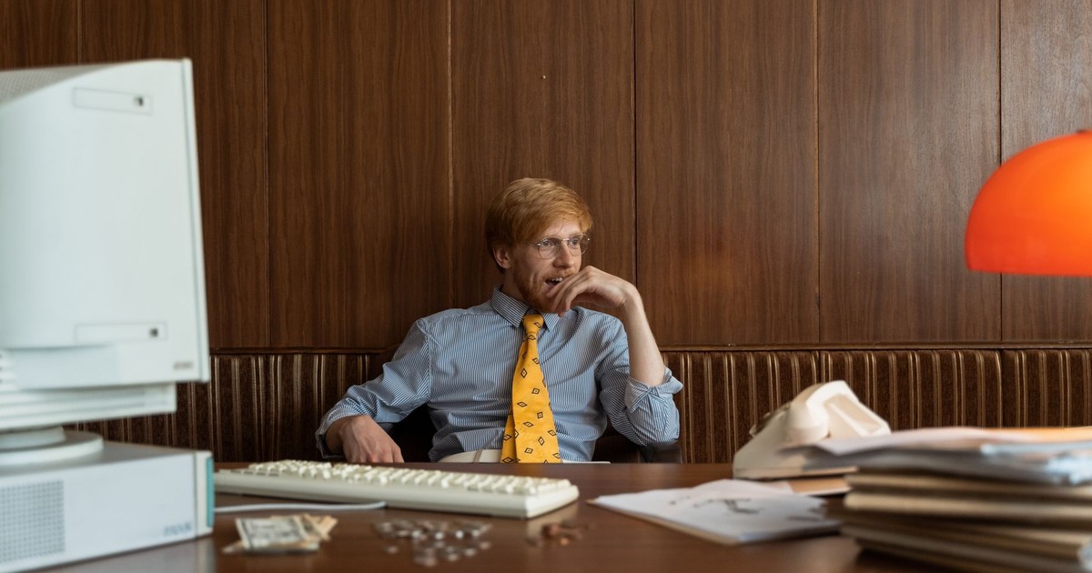 A man in a vintage office setting, looking contemplative with old computer and files.