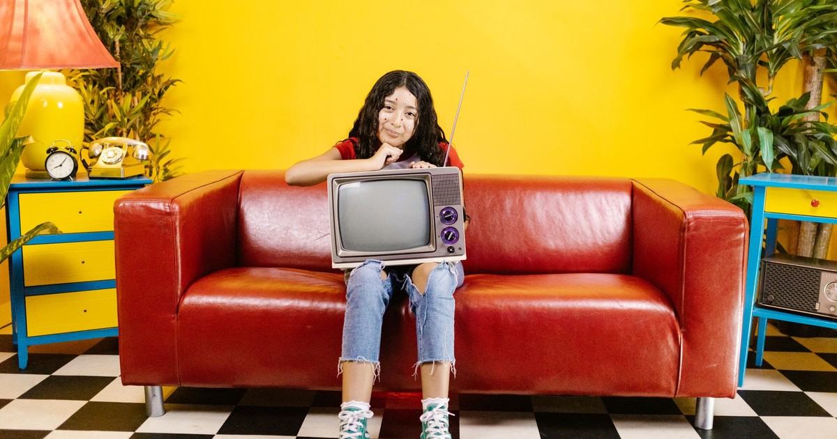 A young woman with a CRT TV sits on a red couch in a colorful living room.