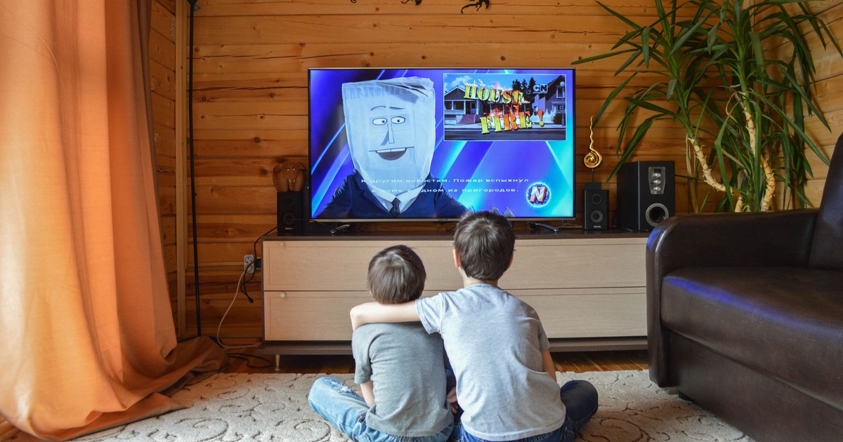 Back view of faceless preteen boy in casual clothes hugging little brother while sitting together on floor and watching TV