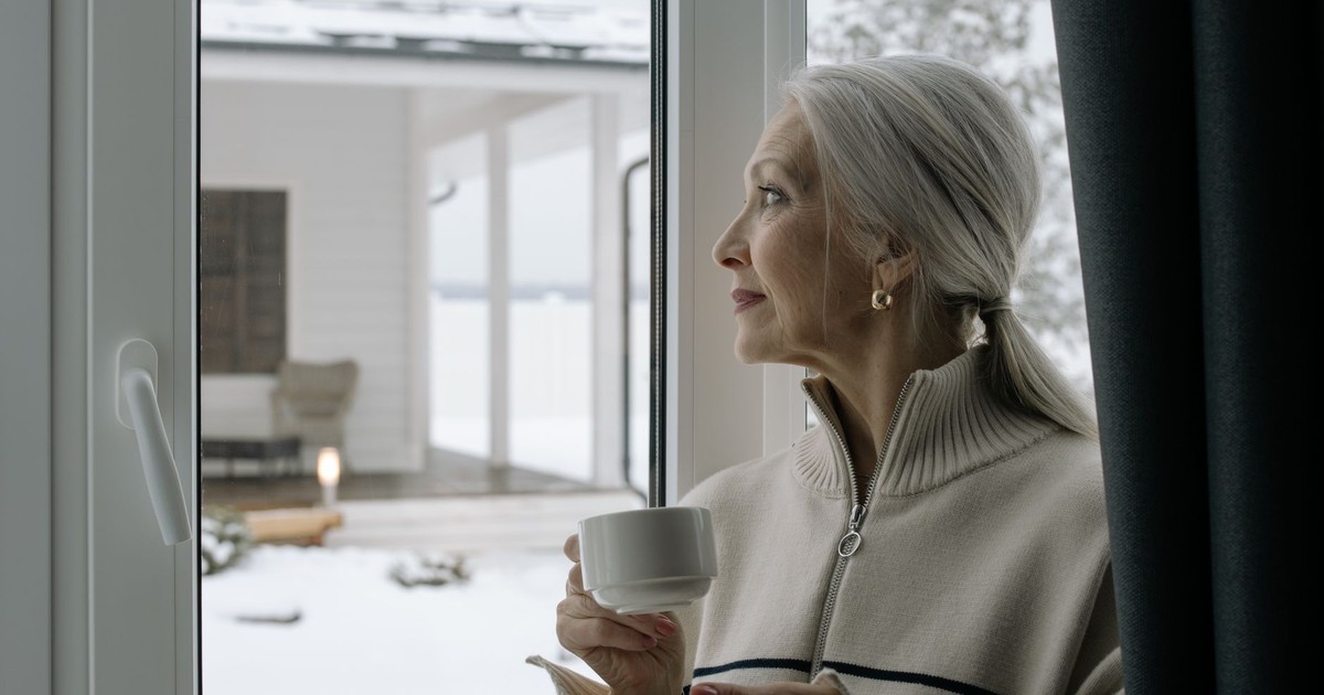 Elegant senior woman with gray hair holds a coffee cup, gazing out a window during winter.