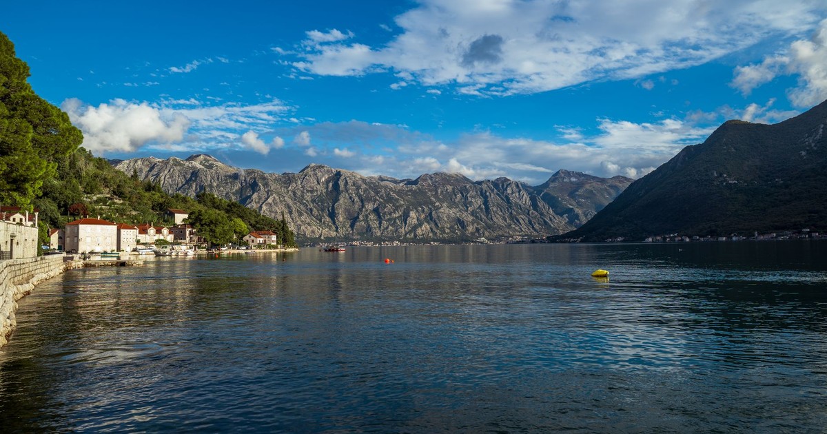 Scenic view of a peaceful coastal town with mountains along a tranquil lake under a bright blue sky.