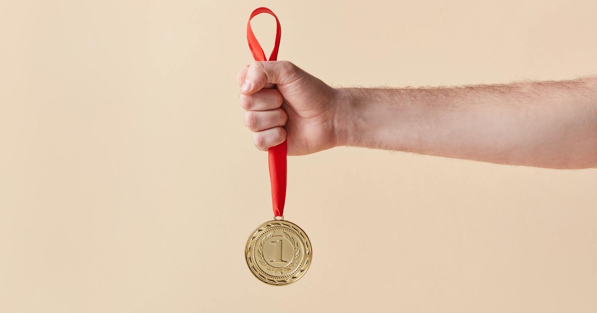 A hand proudly holds a gold medal with a red ribbon on a beige background.