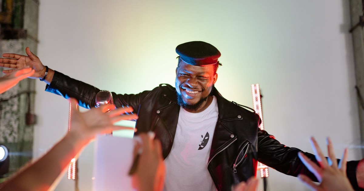 Smiling African American performer in a black leather jacket engaging with an audience during a lively indoor concert.