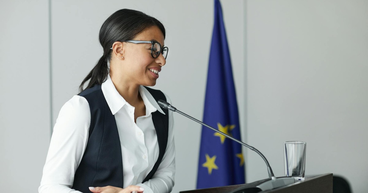 Confident woman speaker at podium with EU flag, delivering a speech indoors.