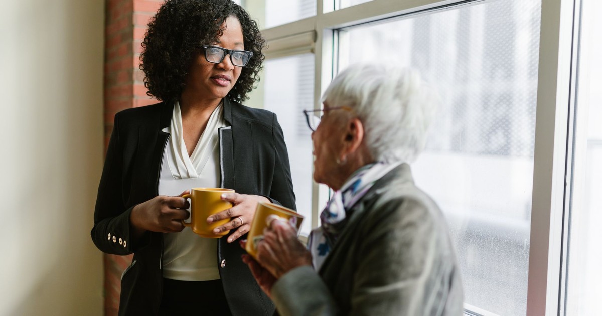 Two women in business attire chatting by a window with coffee mugs in an office setting.