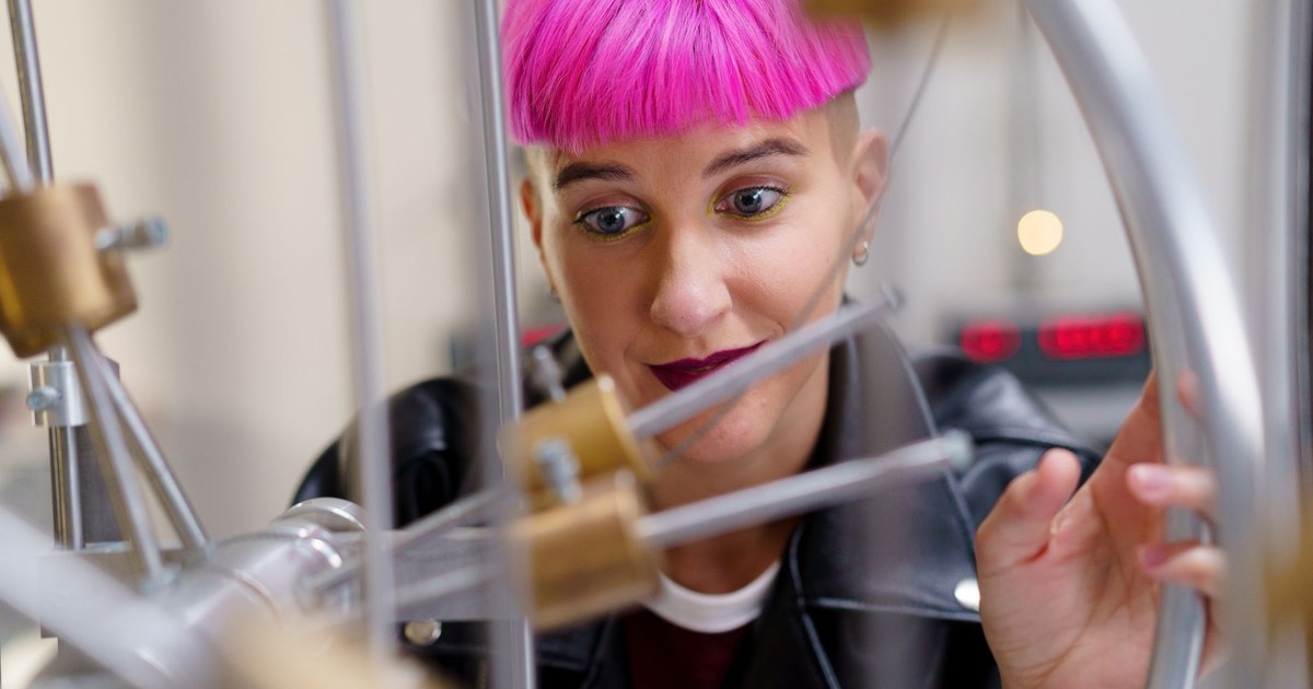 A curious young woman with pink hair examines scientific equipment in a lab setting.