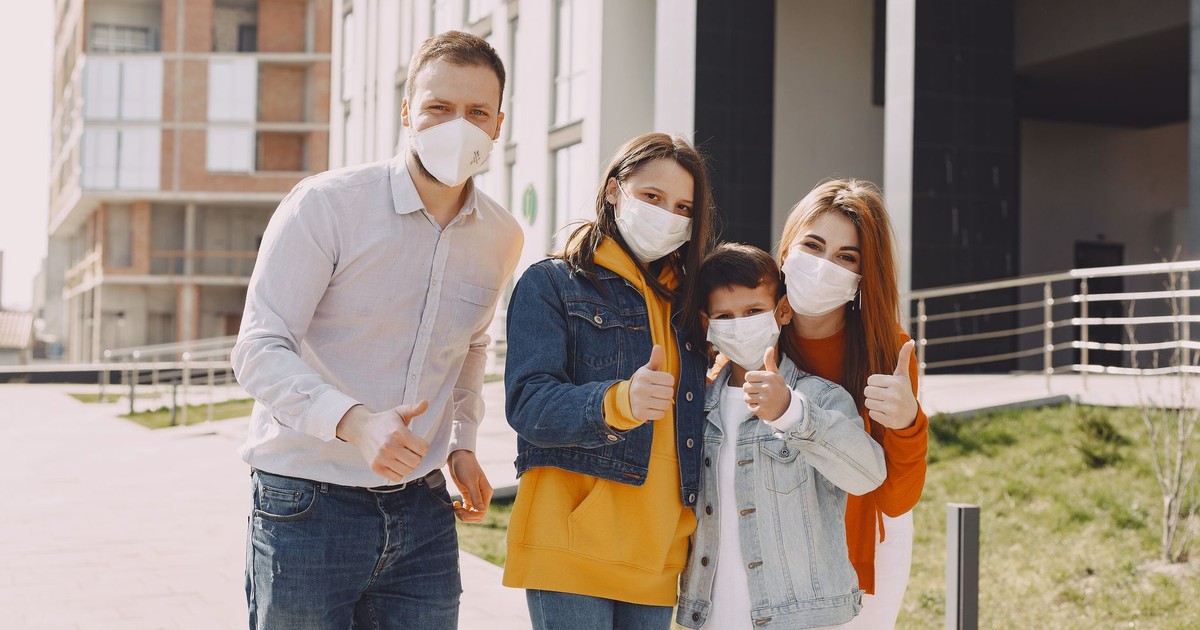 Optimistic family in protective masks and casual clothes standing together on street and showing thumbs up gesture while looking at camera