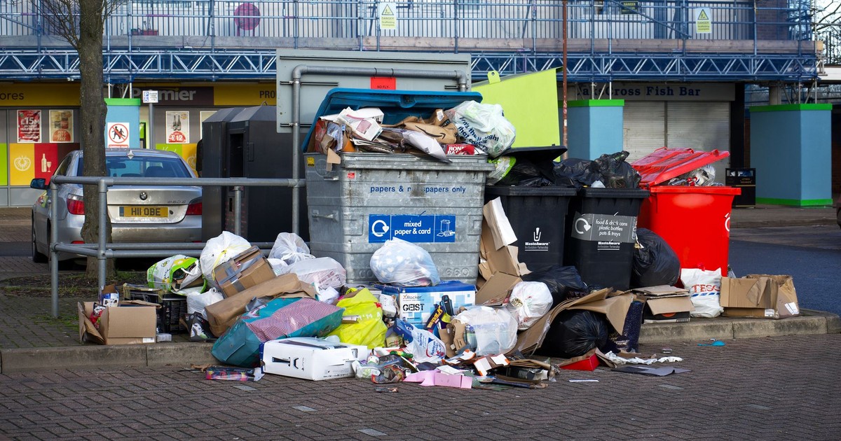 An urban scene depicting overflowing bins and garbage in a parking lot, indicating waste management issues.