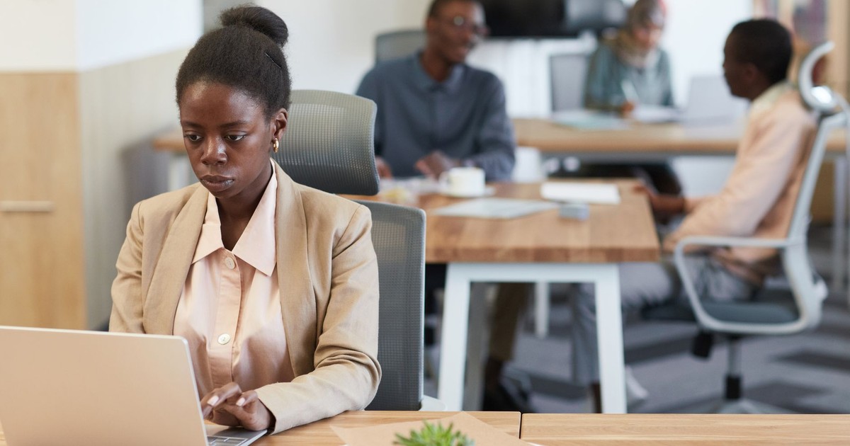 Young African woman in a blazer working intently at a laptop in a bustling office environment.
