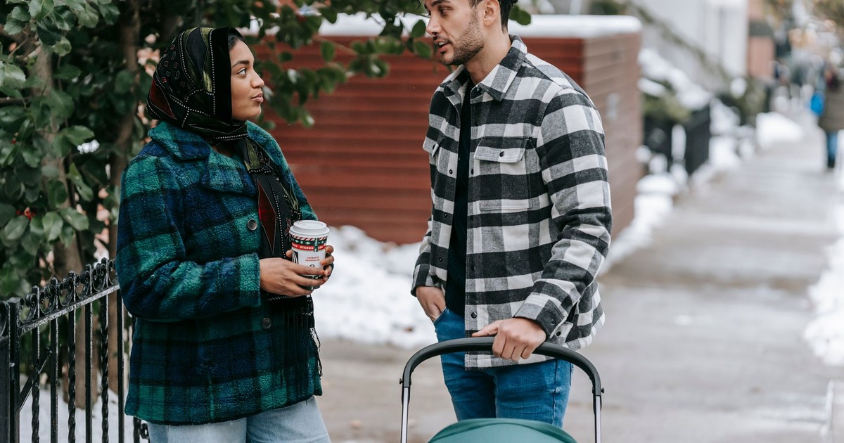 Ethnic woman wearing headscarf standing with cup of takeaway drink and talking to husband standing near stroller on snowy street