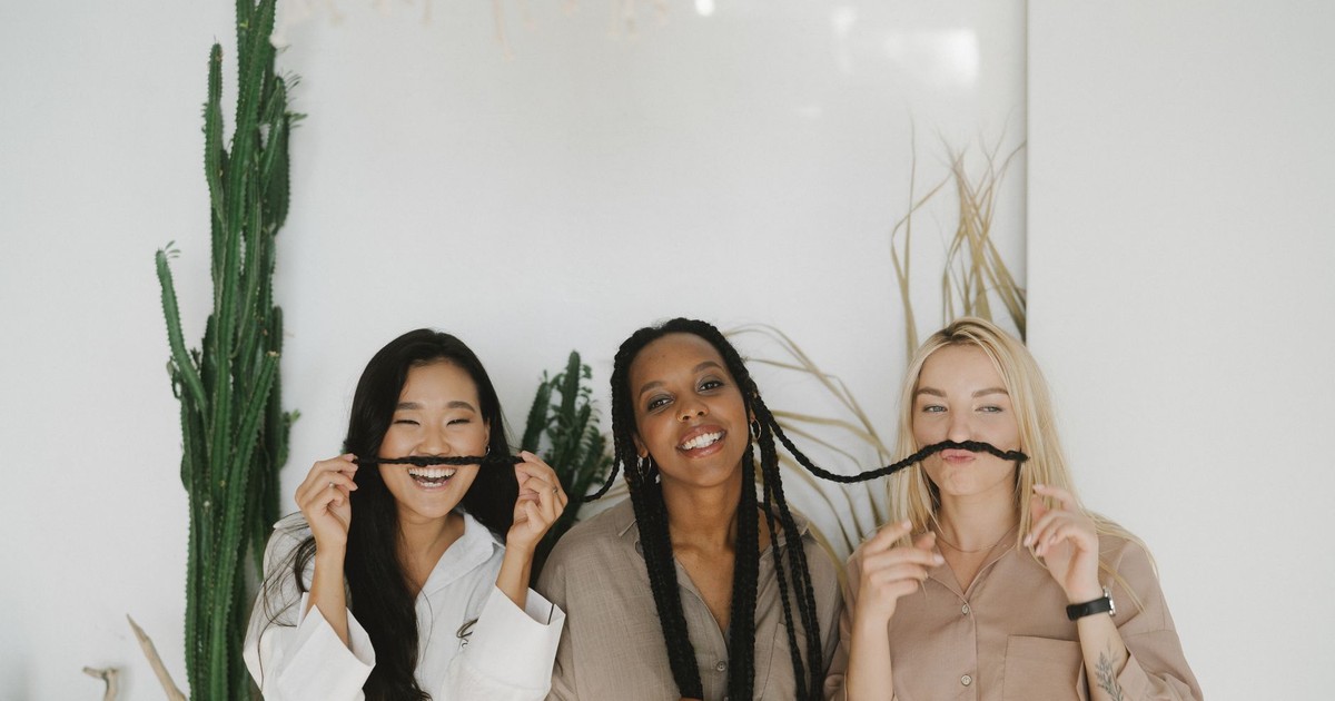 Three women playfully posing with hair mustaches, enjoying a light-hearted moment together indoors.