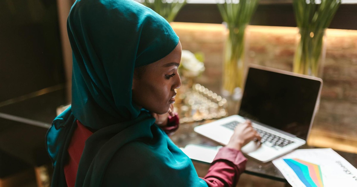 A Muslim woman in a teal hijab works on her laptop at a home office with flower arrangements.