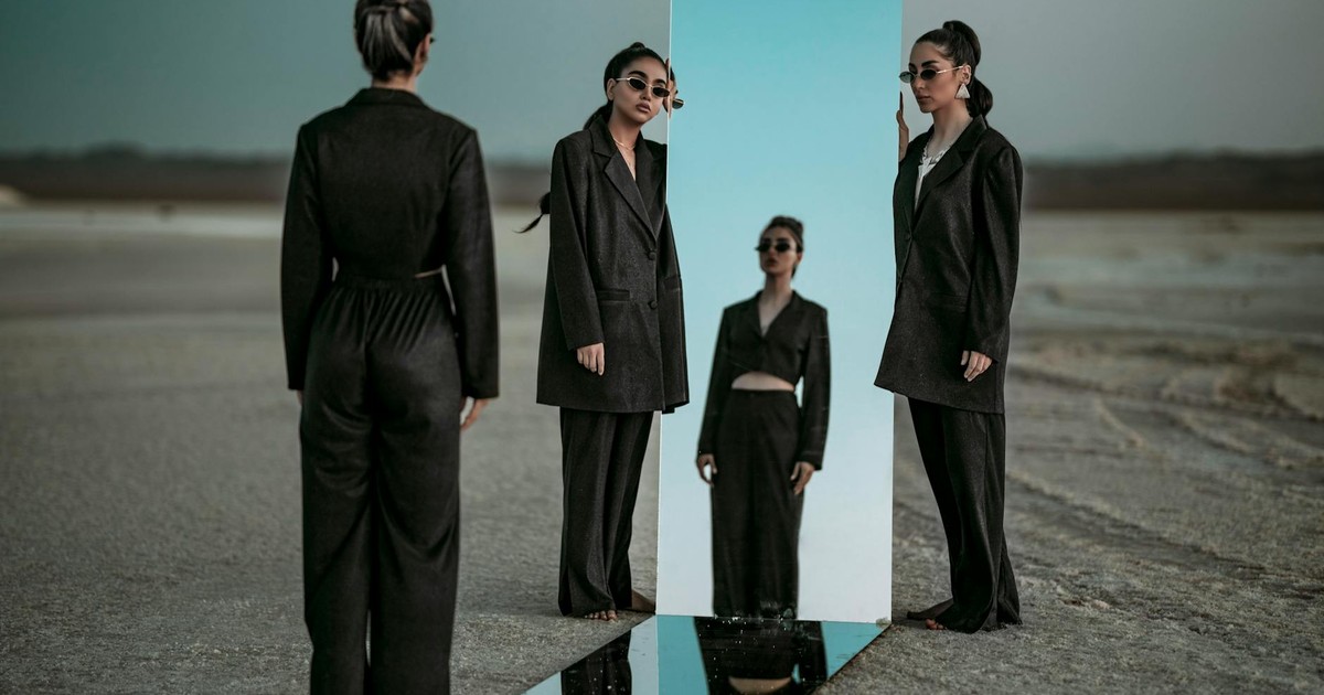 Elegant woman in black clothing reflects in a mirror placed outdoors, creating a surreal fashion scene.