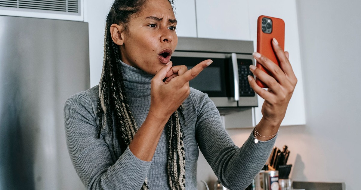 Angry young African American female with long braids in casual clothes pointing at screen nervously while disputing during video call on mobile phone in kitchen