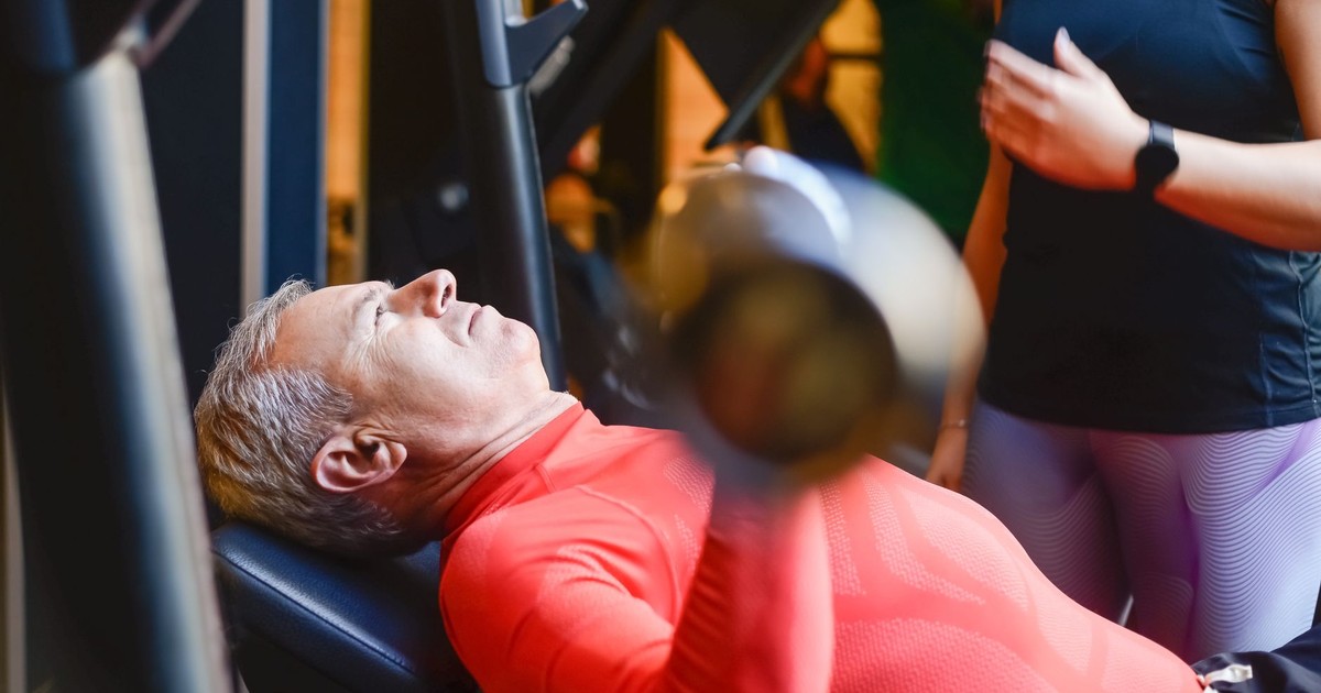 Elderly man exercising with a barbell at the gym under supervision.