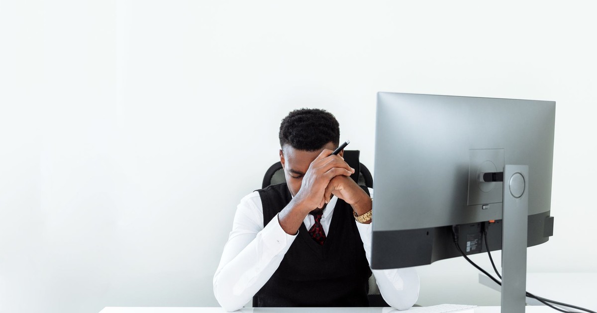 A businessman sitting at a desk, appearing stressed while working at a computer in a bright office.