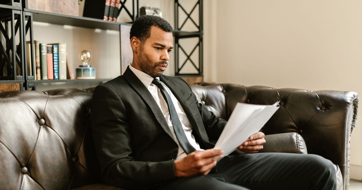 Businessman in suit reading papers on a leather sofa in a modern office setting.