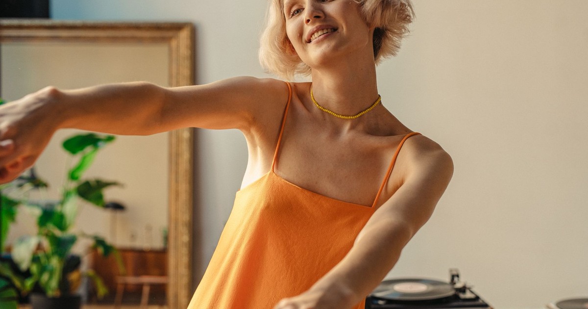 A cheerful woman in an orange spaghetti strap dress dancing joyfully indoors with a smile.
