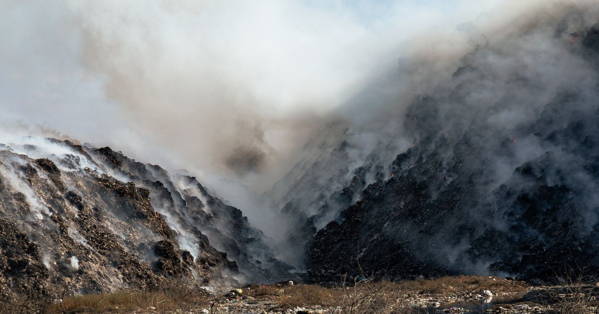 Thick smoke billows from a burning landfill surrounded by piles of waste and trash.