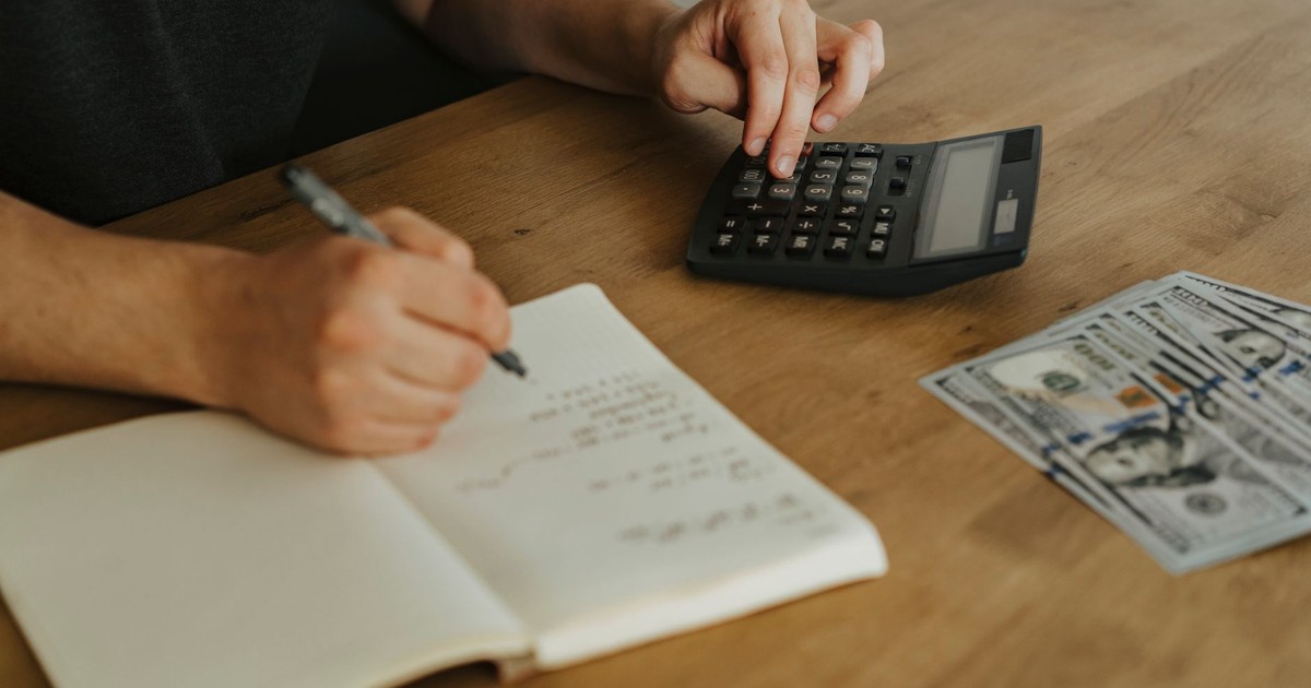 Close-up of hands using a calculator with notebook and cash on a wooden table.