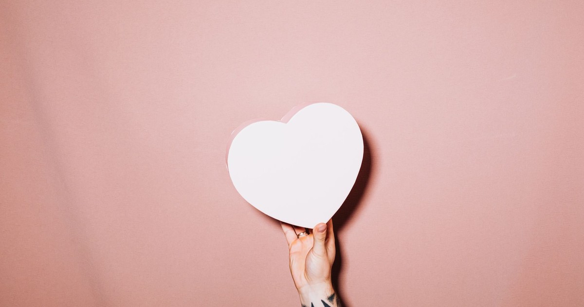 A tattooed hand holds a white heart shape against a pink minimalist background.