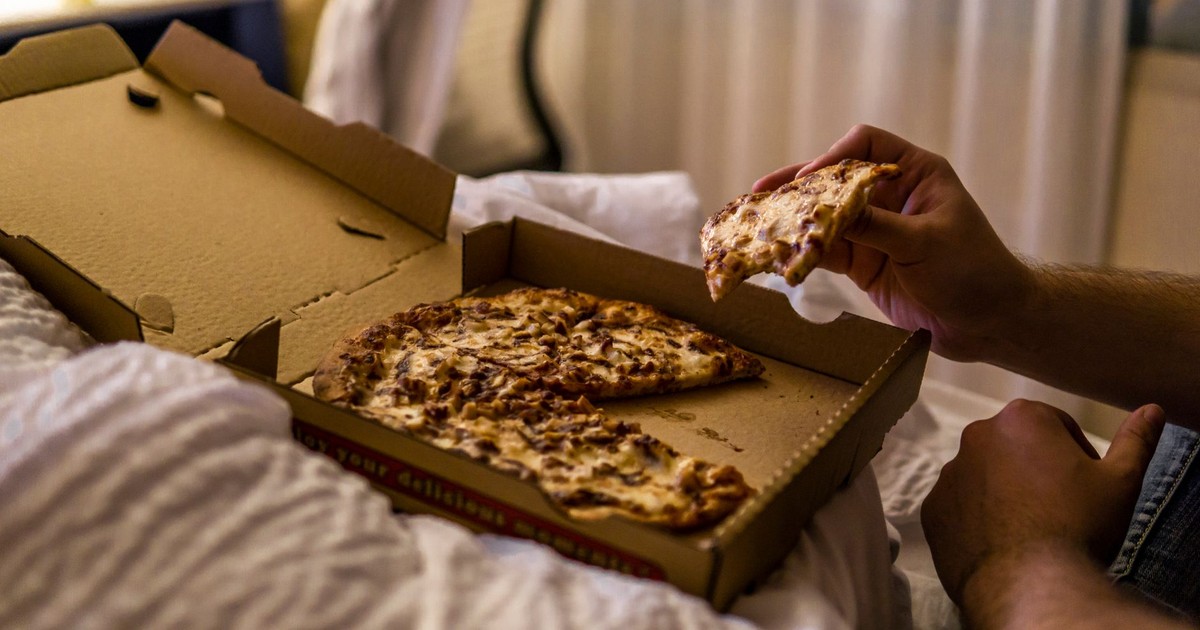 From above side view of crop anonymous male eating tasty slice of pizza with cheese in takeaway cardboard box on crumpled blanket in bed in apartment