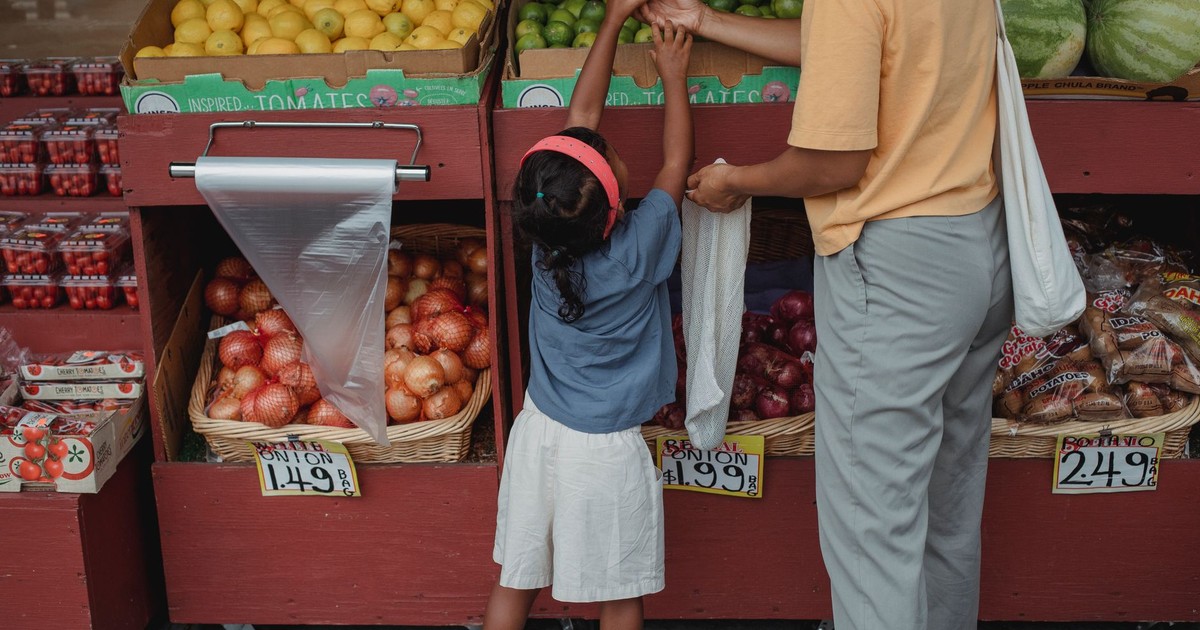 Back view of anonymous ethnic small girl choosing limes from box standing near mother with reusable bag