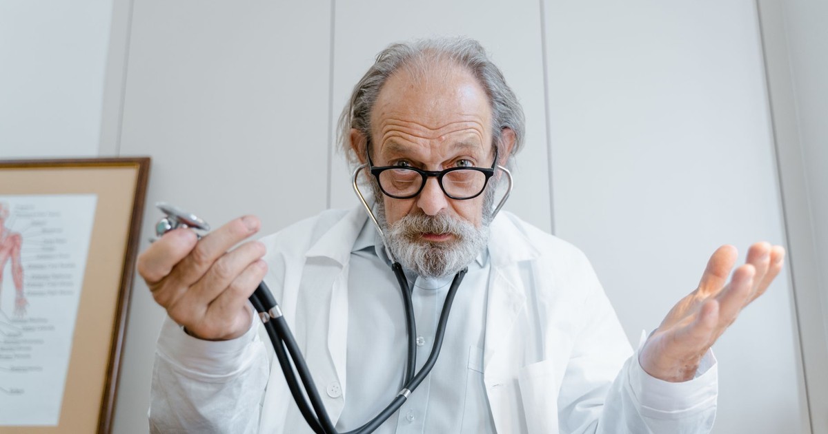 Senior doctor gesturing with stethoscope in a clinical setting, emphasizing telemedicine.