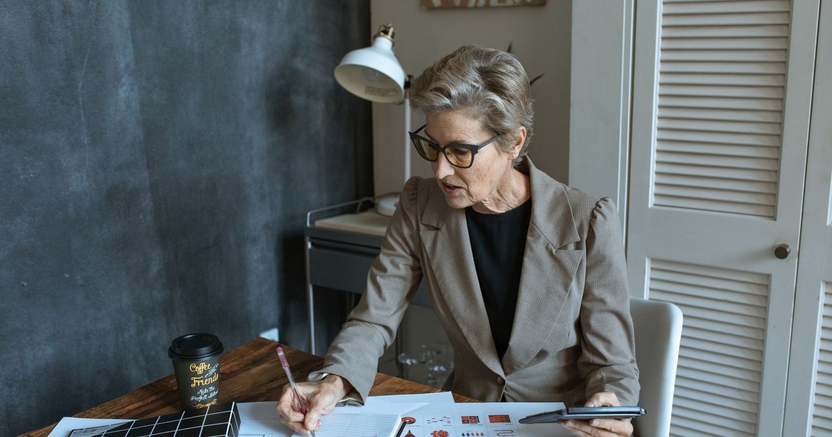 From above of concentrated senior female manager in formal outfit and eyeglasses taking notes in planner while working at table with various documents and takeaway coffee in cozy office