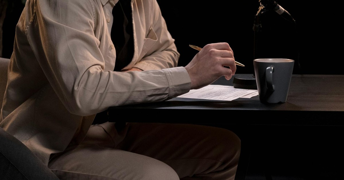 A man in a studio setting working on a podcast with microphone, pen, and coffee cup.