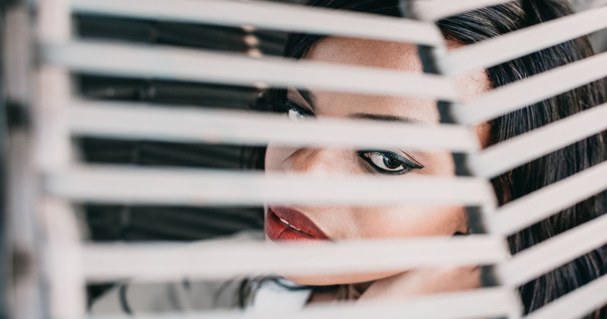 Close-up of a stylish woman peering through window blinds indoors.