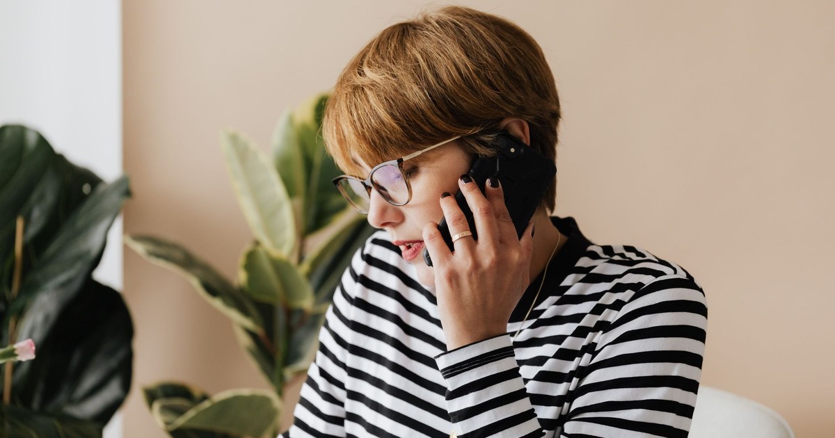 Busy short haired adult lady in eyeglasses sitting in comfortable armchair and talking on smartphone while working in modern office
