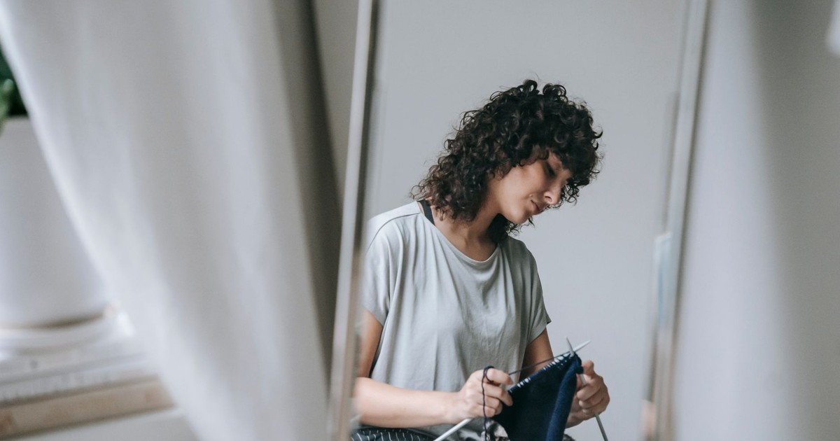 Side view of content young ethnic female with curly hair knitting warm cloth while sitting near mirror in modern apartment