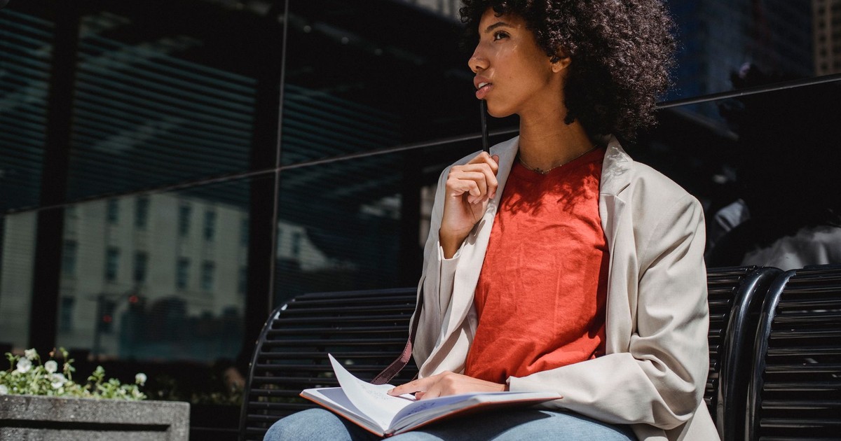 Woman sits outdoors on a bench, writing in a notebook in a city environment.