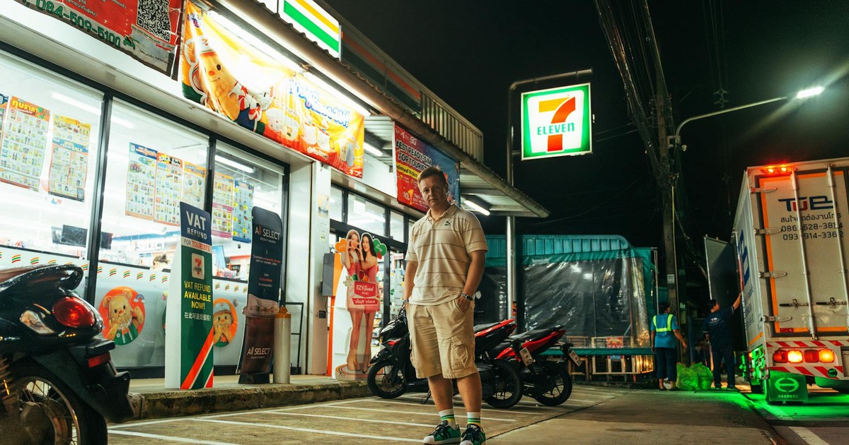 A man stands outside a brightly lit 7-Eleven store at night in Phuket, Thailand.
