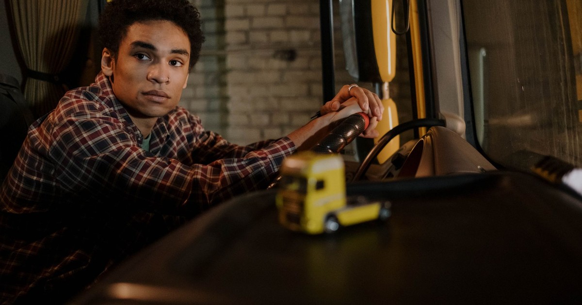 A young man sits in the cabin of a truck, hands on the steering wheel, with a model truck on the dashboard.
