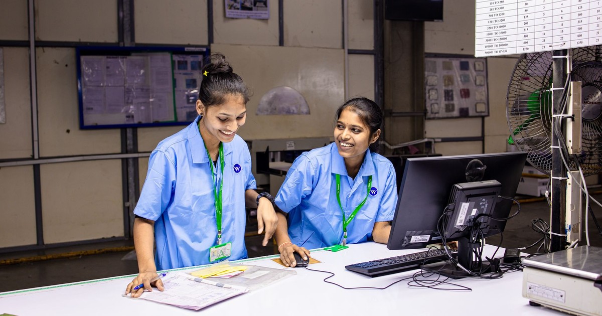 Two female workers discussing tasks at a factory workbench, smiling and engaged.