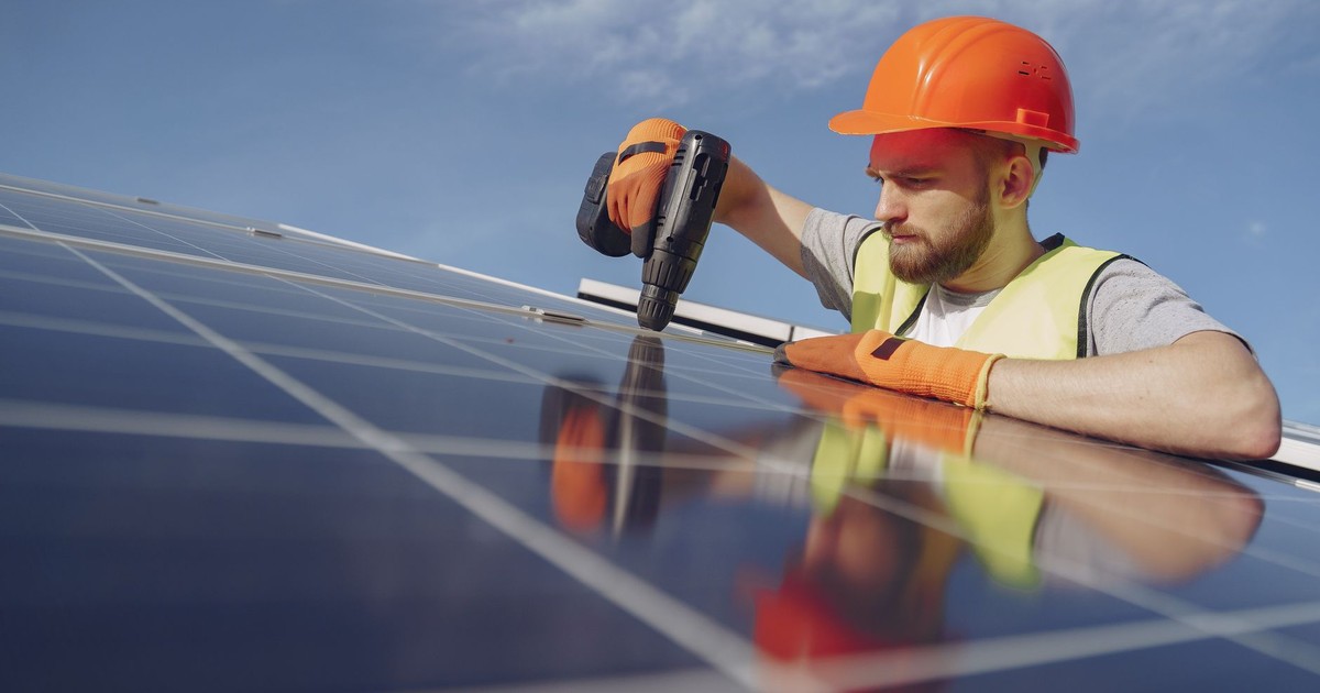 Low angle of serious bearded male electrician in hardhat and protective gloves installing solar photovoltaic panel system using drill