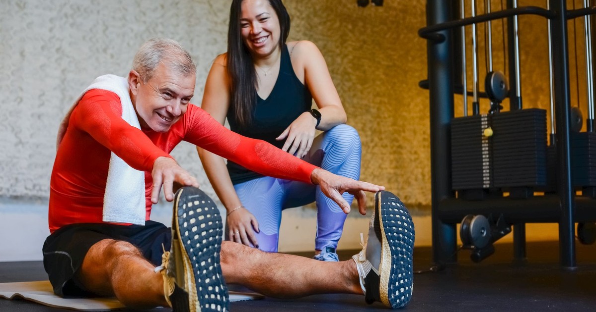 Elderly man in a gym stretching with a fitness coach, promoting a healthy lifestyle.