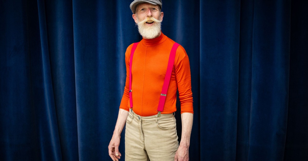 Portrait of an elderly man wearing orange shirt and suspenders against blue curtain.