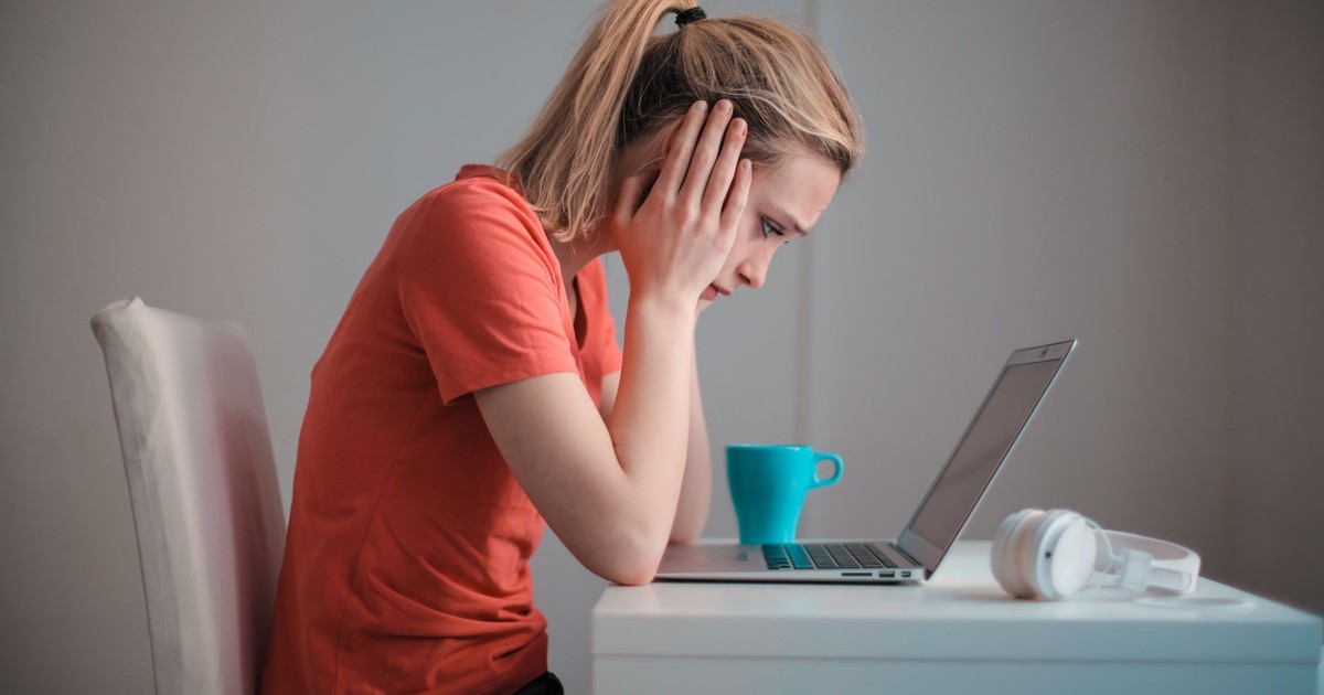 Side view of thoughtful worried female student in casual clothes focusing on screen and trying to solve problem while sitting at table with blue mug of hot drink and headphones and working on laptop at home