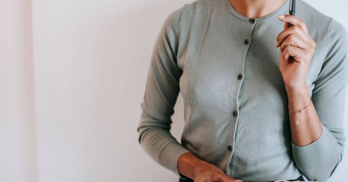 Crop anonymous female consultant wearing formal clothes standing with clipboard against white wall and touching chin with pen in contemplation
