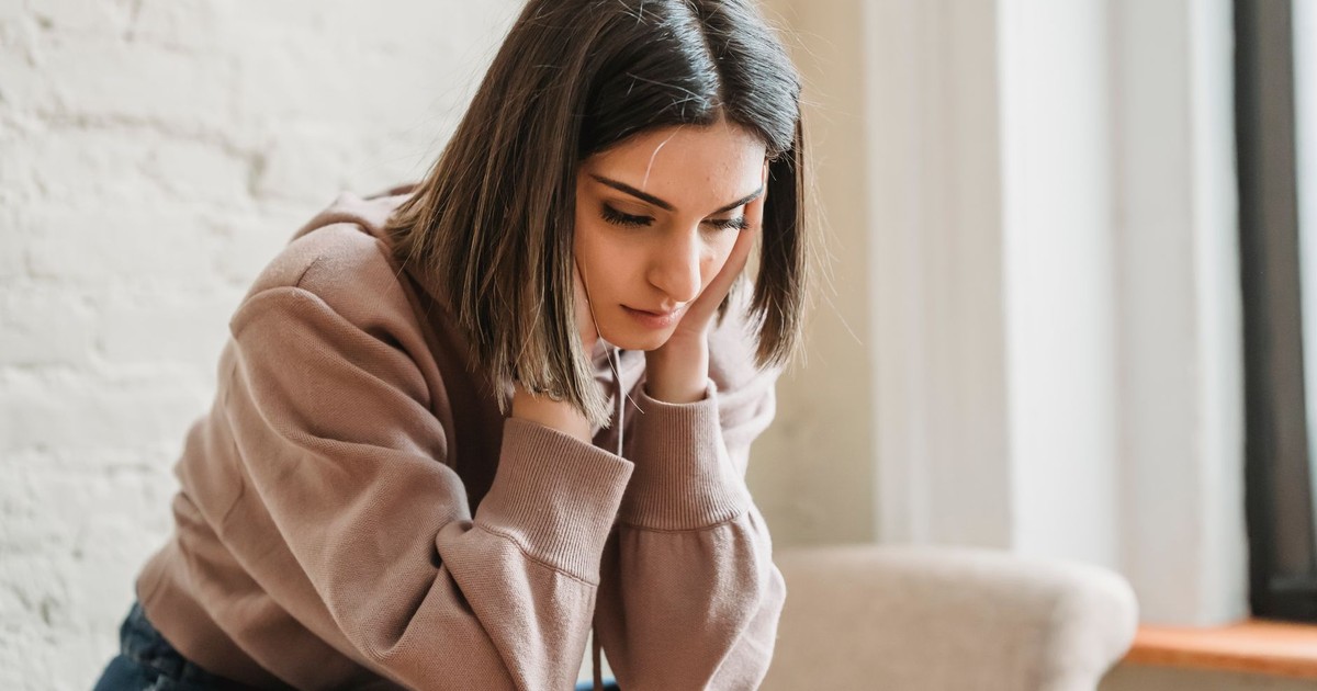 Unhappy female in casual wear touching face and looking down while sitting on sofa in light living room at home