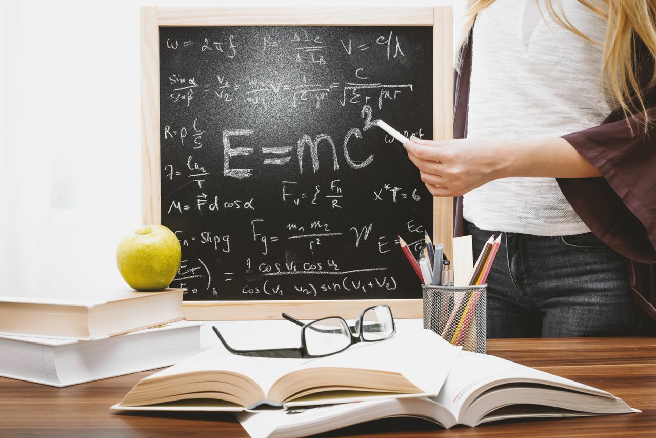Woman writing physics equations on a blackboard with books and an apple on the desk