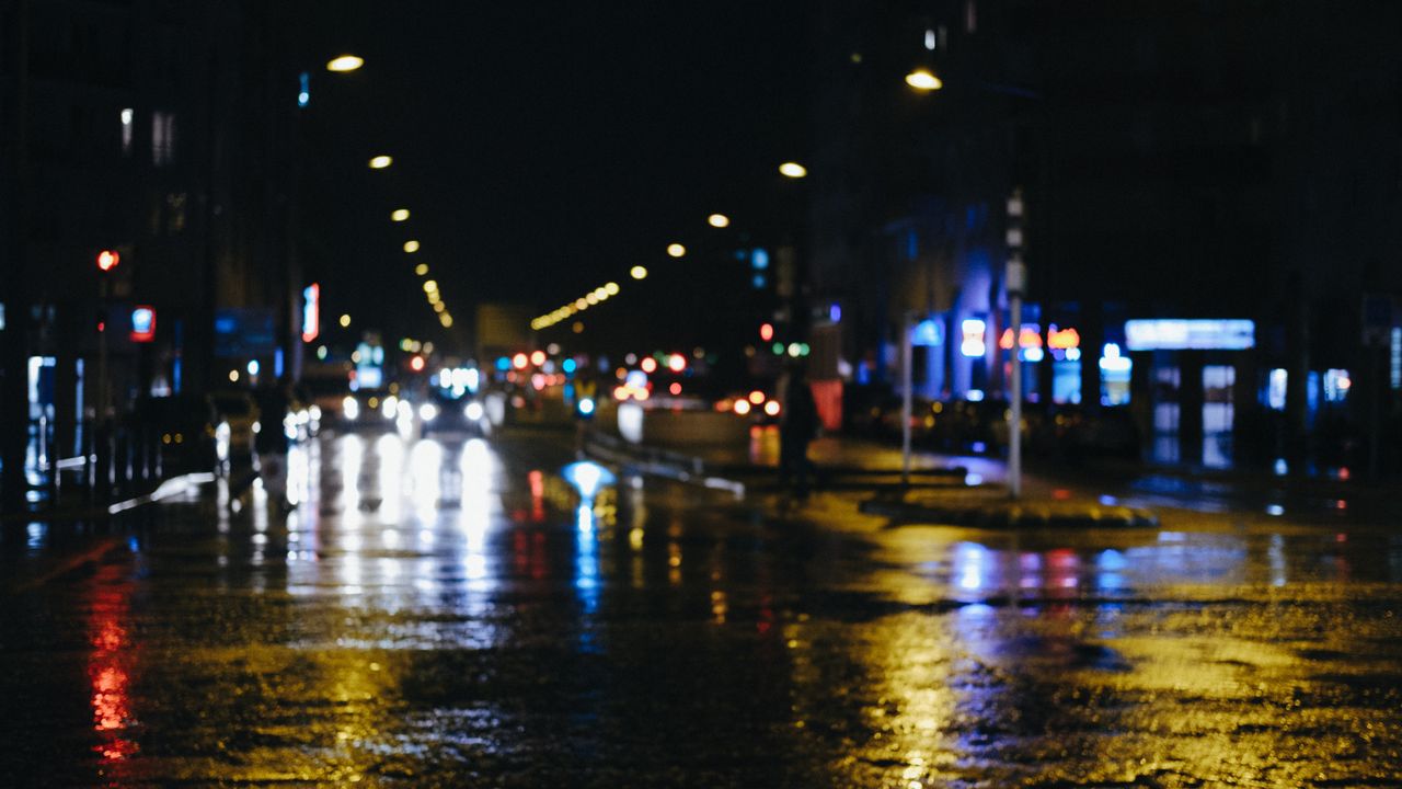 Blurred urban street at night with reflections of city lights on wet pavement.