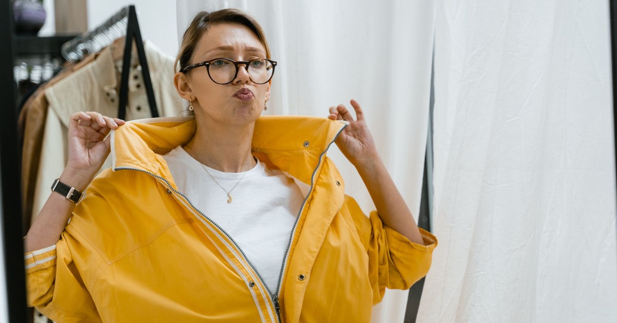 Woman playfully trying on a vibrant yellow jacket in a clothing store dressing area.
