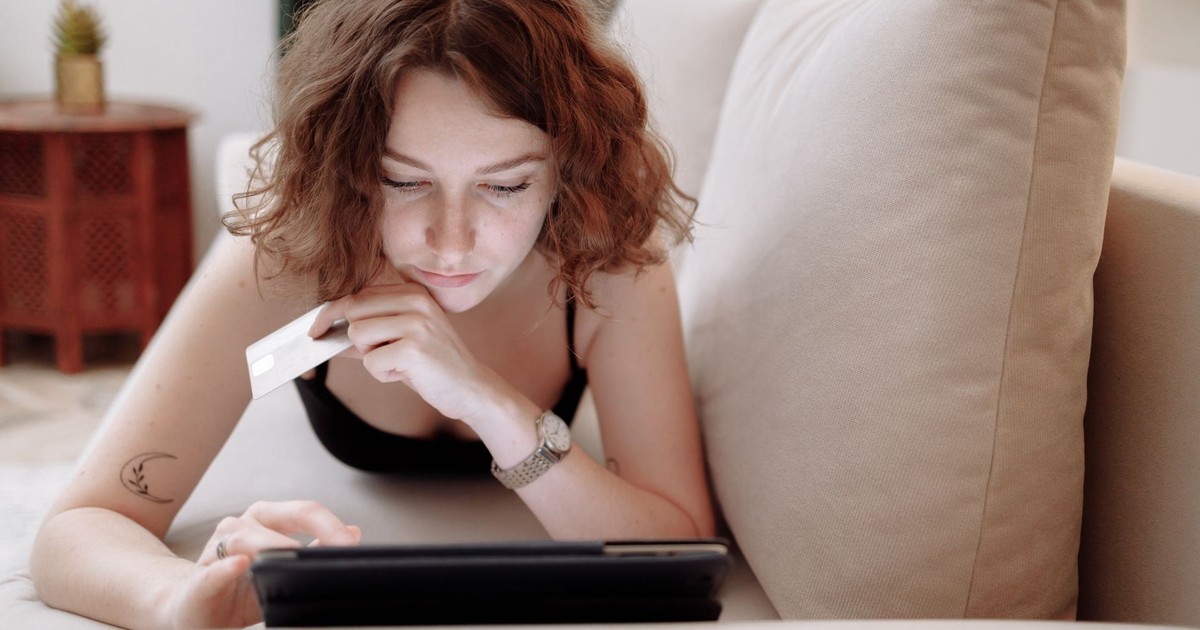 Young woman using a tablet and credit card for online shopping, lying on a couch indoors.
