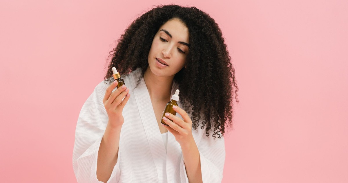 A woman with curly hair in a white robe examines two cosmetic oil bottles against a pink background.