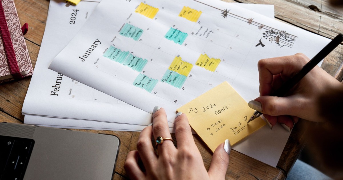 Close-up of a person writing new year's goals on a sticky note with a 2024 calendar in the background.